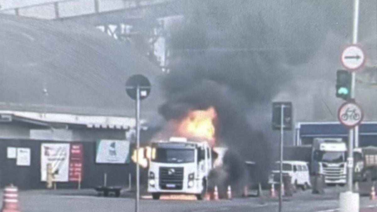 Caminhão Santos carregado com piche pega fogo na via, com chamas saindo da parte frontal e fumaça ao redor, em Santos, e deixa motorista ferido.