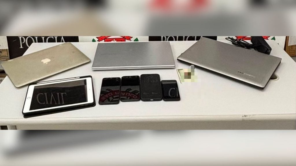 Mesa de evidências com aparelhos celulares e documentos apreendidos em operação policial, com lona “POLICIA” ao fundo, em Santos, relacionada a suspeita de crimes sexuais contra mulheres e adolescentes.