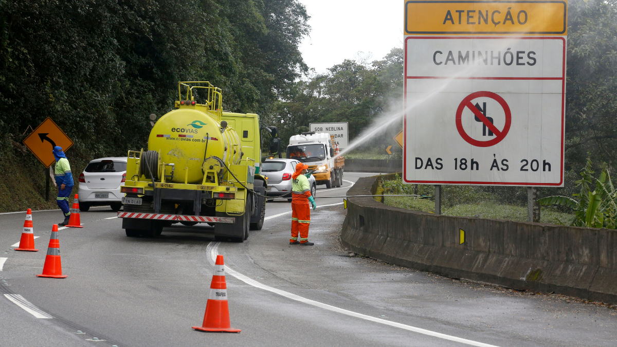 Trânsito em obra com caminhão de manutenção e cones na via, com placa de atenção indicando restrição das 18h às 20h na região de Anchieta e Imigrantes nesta semana.