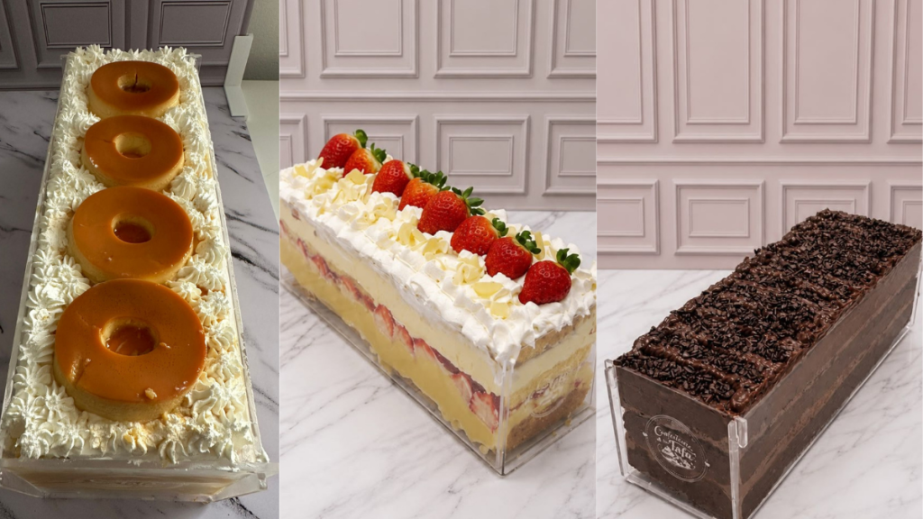 Três opções de bolo decorado em bancada de mármore: bolo com cobertura clara e letras/figuras douradas, bolo retangular com morangos e chantilly e outro bolo retangular de chocolate com farelos, em composição de vitrine para o Festival de Fatias em Praia Grande.