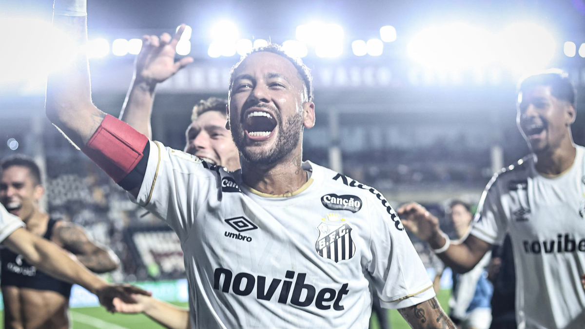 Neymar comemora com sorriso aberto e gesto com o braço levantado em partida de futebol no estádio, enquanto jogadores ao fundo celebram. Neymar ironiza rivalidade com Thiago Mendes: “Tenho coisa melhor pra resolver”.
