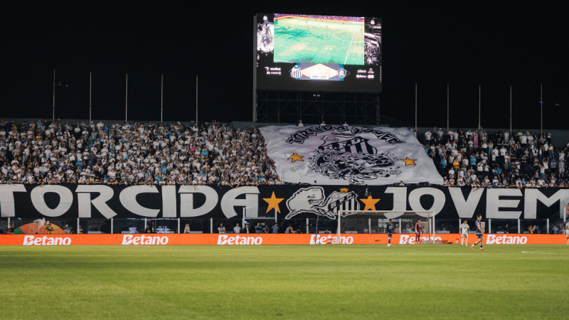 Estádio com torcida lotada e faixa de Santos no gramado durante jogo do Santos x Mirassol, com painel eletrônico e jogadores em campo ao fundo.