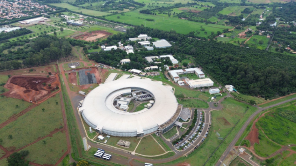 cnpem campinas: Vista aérea de instalações do CNPEM em Campinas, mostrando pavilhões, áreas verdes e um grande prédio circular, com referência às Bolsas de Verão do CNPEM com inscrições abertas.