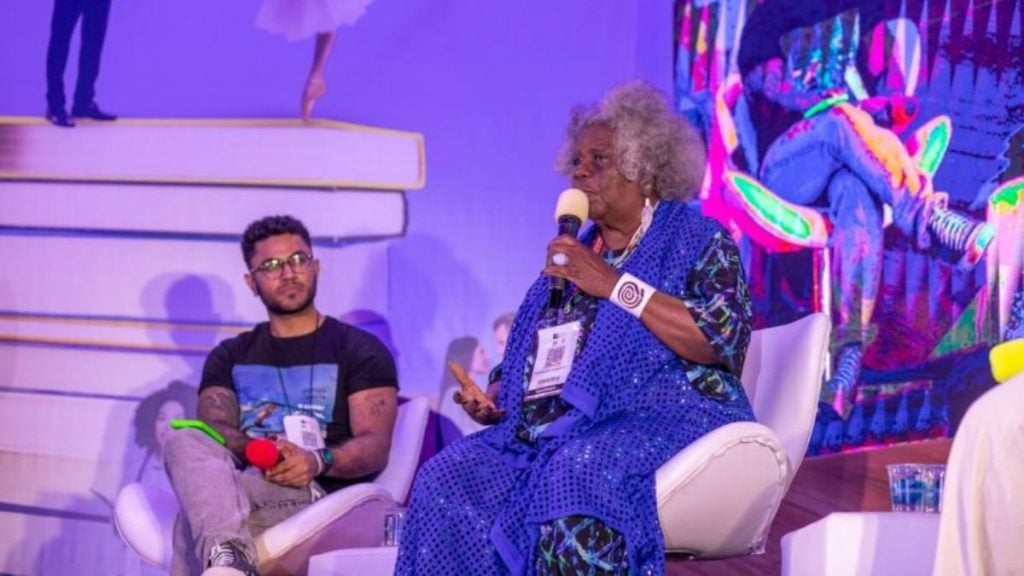 Bienal do Livro de São Paulo 2026: Conceição Evaristo em evento de palco, sentada em uma cadeira e segurando microfone, durante conversa com plateia, ao lado de painel e convidados; Foto: Divulgação.