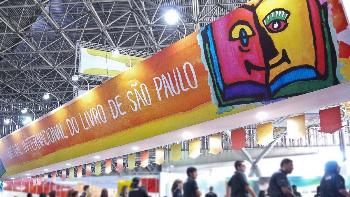 Bienal do Livro de São Paulo 2026: Banner colorido com o texto “BIENAL INTERNACIONAL DO LIVRO DE SÃO PAULO” em área interna de feira, com estrutura metálica do pavilhão e pessoas ao fundo.