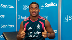 Endrick aparece sorrindo em coletiva de imprensa com colete do Real Madrid, no fundo há painéis azuis com o patrocinador Sanitas. A imagem indica interesse do Liverpool.