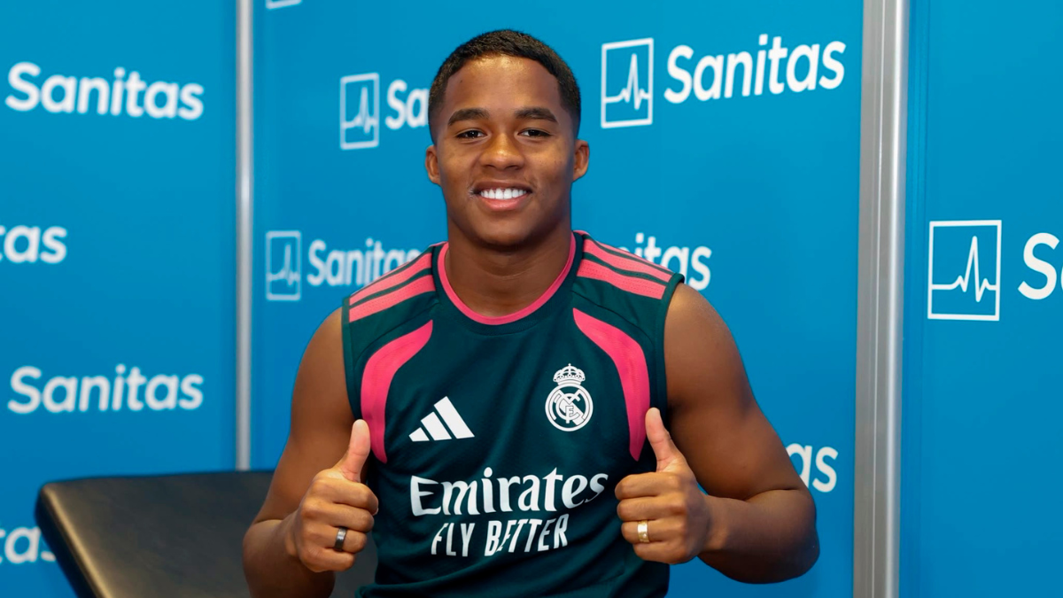 Endrick aparece sorrindo em coletiva de imprensa com colete do Real Madrid, no fundo há painéis azuis com o patrocinador Sanitas. A imagem indica interesse do Liverpool.