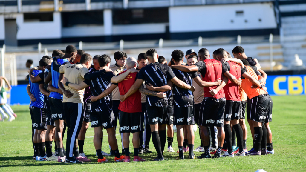Elenco da Ponte Preta reunido em campo, durante atividade de equipe em estádio, com jogadores agrupados e uniformes azuis, vermelhos e pretos.