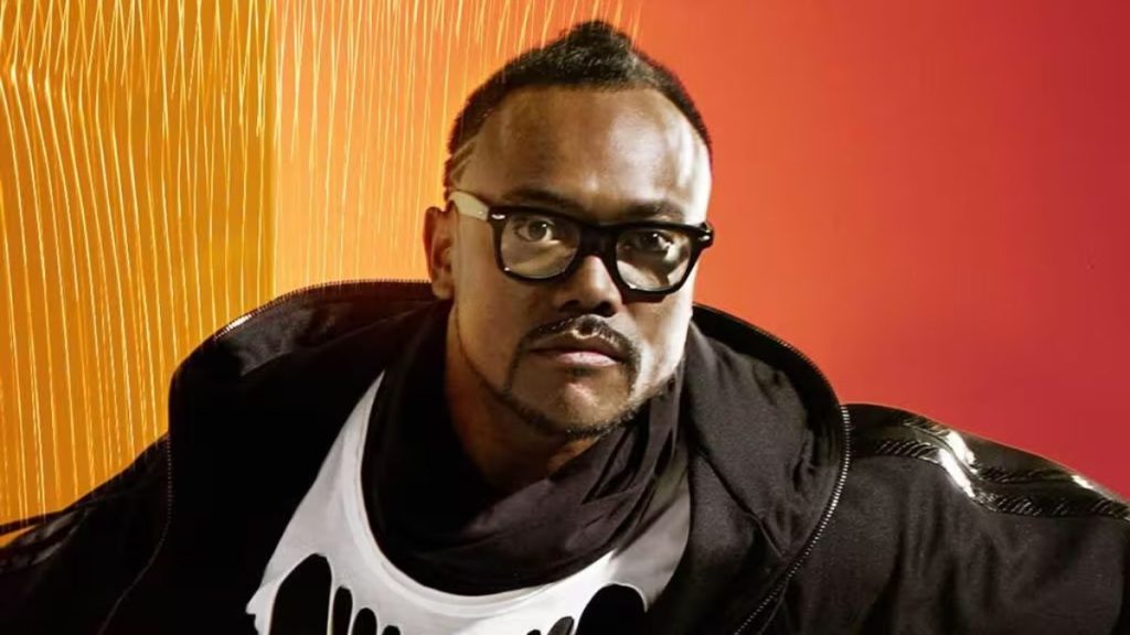 Rock in Rio 2026: Black Eyed Peas em destaque: foto do integrante do grupo com óculos escuros, expressão séria, usando jaqueta e camisa, ao fundo com textura laranja e vermelha do show.