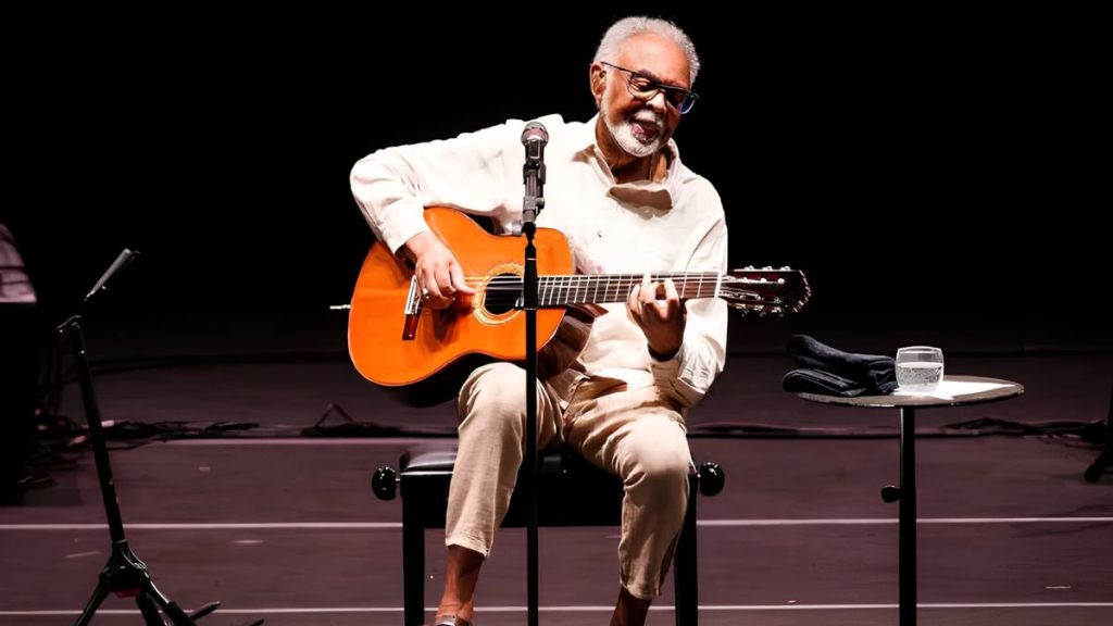 Rock in Rio 2026: Gilberto Gil se apresenta no palco com violão, sentado em um banquinho, usando camisa clara e barba branca, em foto de reprodução do festival.