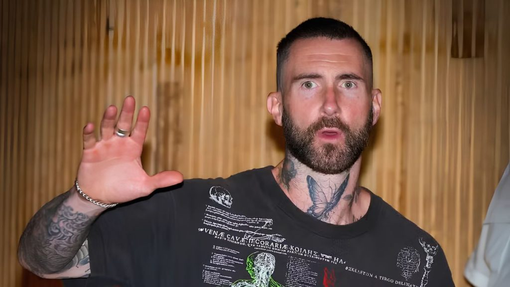 Maroon 5 em destaque no Rock in Rio 2026: cantor com tatuagens e barba, de camisa preta, fazendo gesto de aceno em foto de divulgação do festival, com apresentação no dia 12 de setembro.
