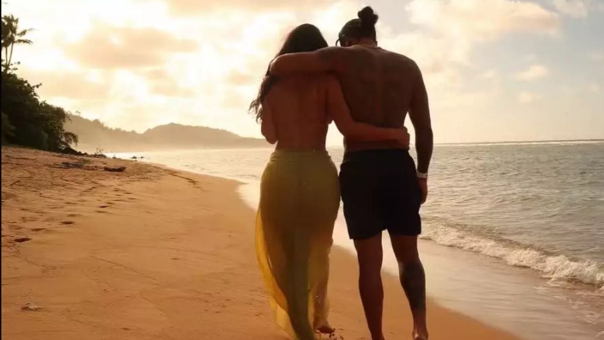 Kim Kardashian e Lewis Hamilton caminhando lado a lado na praia ao pôr do sol, em registros recentes do romance, com o mar ao fundo.