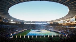 Copa do Mundo Feminina 2027: estádio lotado com campo de futebol visto de arquibancadas, equipes alinhadas e iluminação para a cerimônia de abertura.