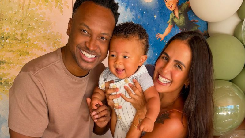 Thiaguinho e Carol Peixinho comemoram 11 meses do filho em uma festa inspirada em livros, em foto sorridente com o bebê no colo, decorada com cenário infantil.