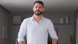 Evaristo Costa em foto promocional, vestindo camisa social branca, falando para a câmera em ambiente interno, com expressão séria ao anunciar conteúdo adulto.