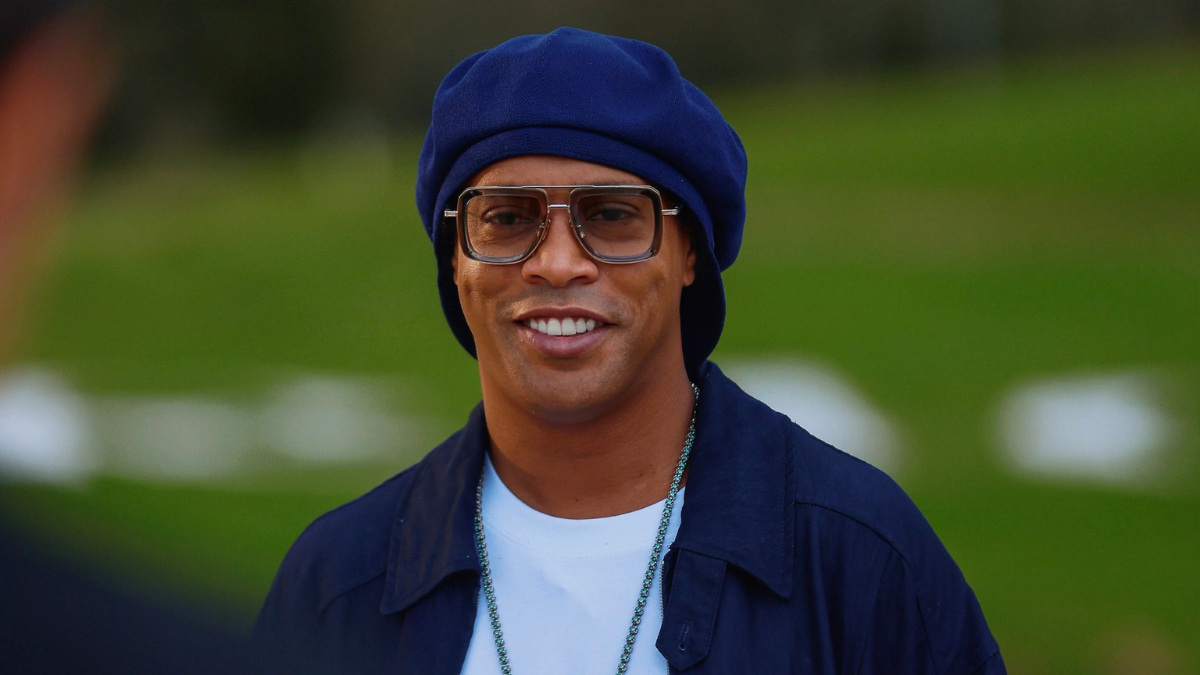 Ronaldinho aparece sorrindo em um estádio, usando boné e óculos, em imagem relacionada ao retorno ao futebol profissional e a possibilidade de atuar na 3ª divisão da Itália.