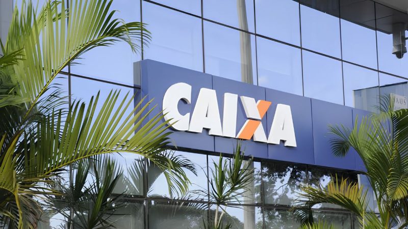 Presos no elevador de uma agência bancária, com sinalização do banco CAIXA na fachada em ambiente com plantas e luz natural no litoral de SP.