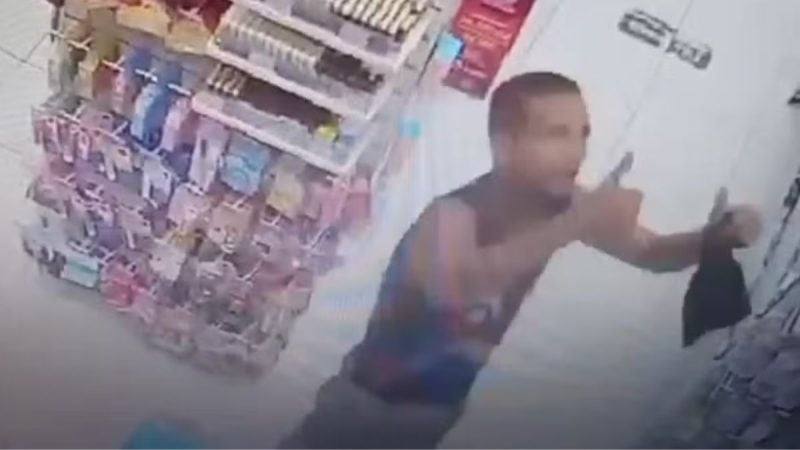 Suspeito acena para câmera antes de roubar farmácia em Valinhos, mostrado em imagem de segurança dentro de uma loja, com homem segurando os braços em gesto de aceno.