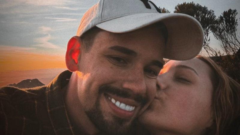 Lucas Borbas: Casal em clima romântico em ambiente externo ao pôr do sol, com um homem sorrindo e abraçando a mulher; fotografia próxima e com chapéu.