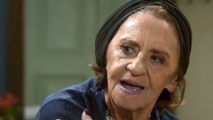 Laura Cardoso: Imagem em close mostra uma mulher idosa, com expressão emocionada, usando um gorro escuro e cabelo curto, em cena de entrevista ou depoimento. Ao fundo há um ambiente interno desfocado.