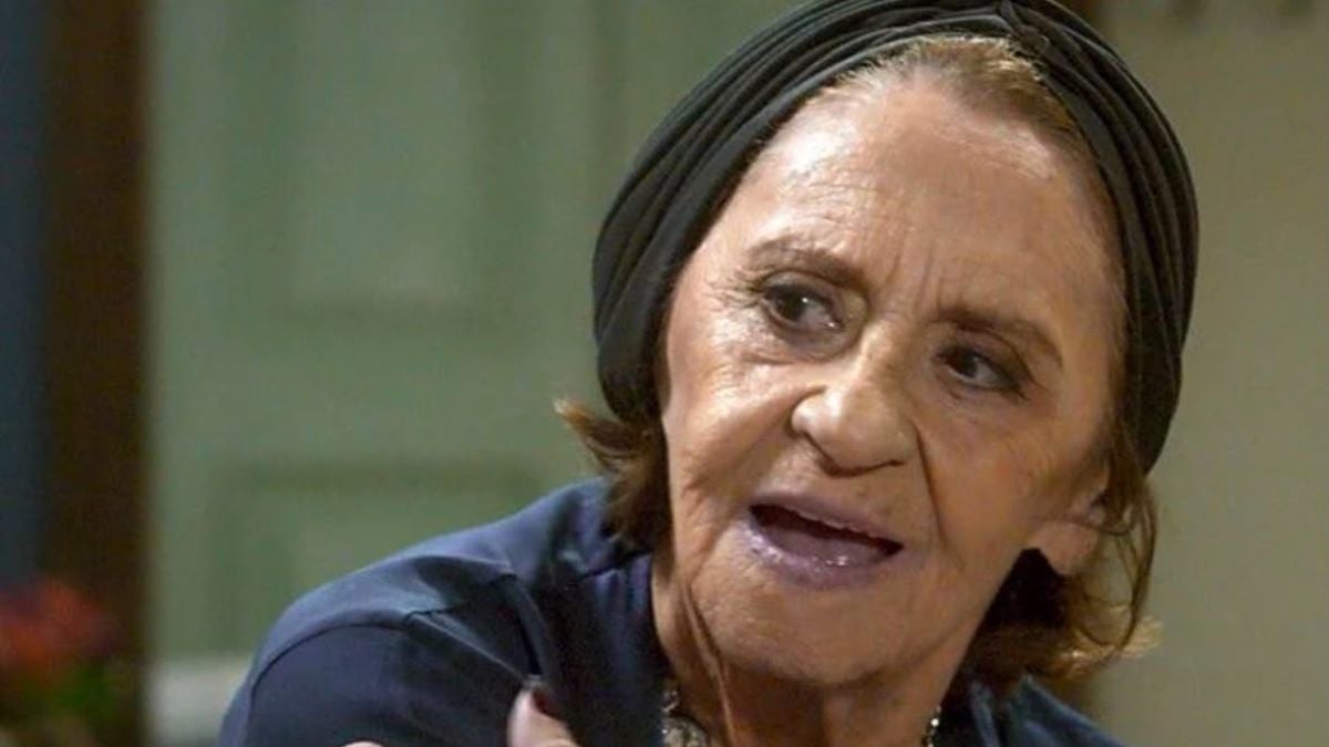 Retrato em close de Laura Cardoso durante entrevista, com expressão séria, usando um lenço na cabeça e fundo desfocado em tom verde.
