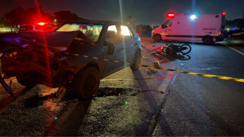 motociclista de 22 anos morre em Cosmópolis: Acidente de trânsito à noite com carro e motocicleta, viatura e ambulância ao fundo; na pista há moto caída e faixas amarelas de isolamento próximo a Cosmópolis.
