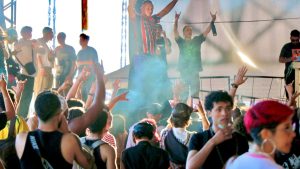 Público em show ao ar livre em Campinas, com pessoas com as mãos para o alto e banda no palco ao fundo durante a Semana da Música em Campinas, com iluminação e fumaça no evento