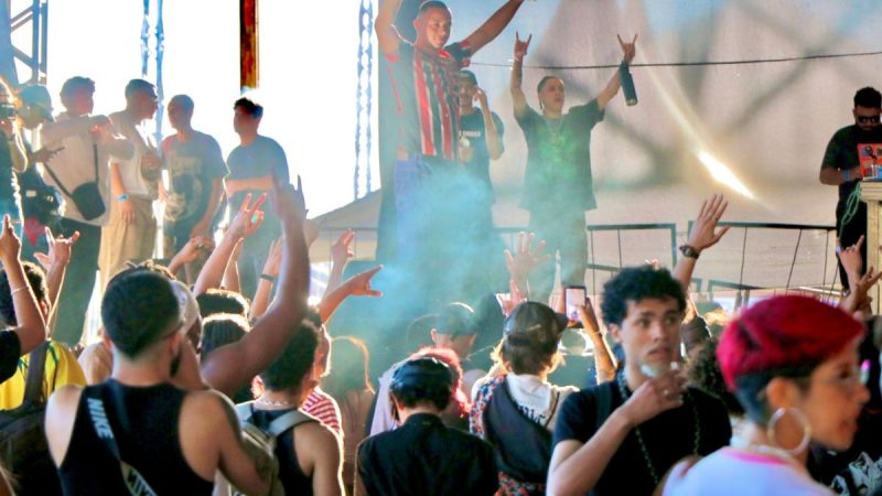Público em show ao ar livre em Campinas, com pessoas com as mãos para o alto e banda no palco ao fundo durante a Semana da Música em Campinas, com iluminação e fumaça no evento