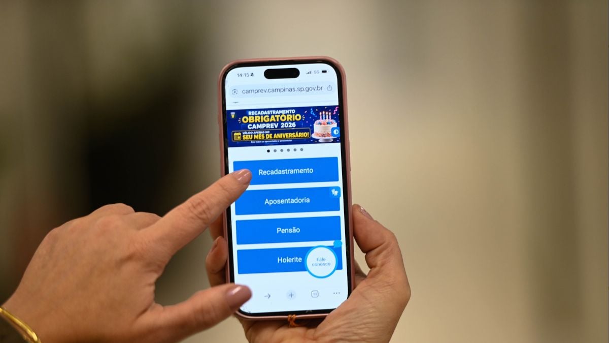 Pessoa usando smartphone com o site do Camprev aberto, na tela aparece o aviso de recadastramento Camprev para beneficiários nascidos entre agosto e dezembro, com menu para Recadastramento, Aposentadoria, Pensão e Holerite.
