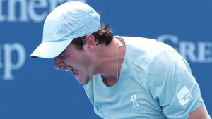 João Fonseca em partida de tênis, curvado e concentrado no saibro, usando boné e camisa azul clara. A imagem faz referência à notícia de que ele está fora do US Open por lesão no abdômen.
