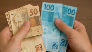 Mãos segurando notas de dinheiro do Brasil, incluindo cédulas de 50 e 100 reais, com foco em reajuste do salário mínimo e valores projetados para 2027.