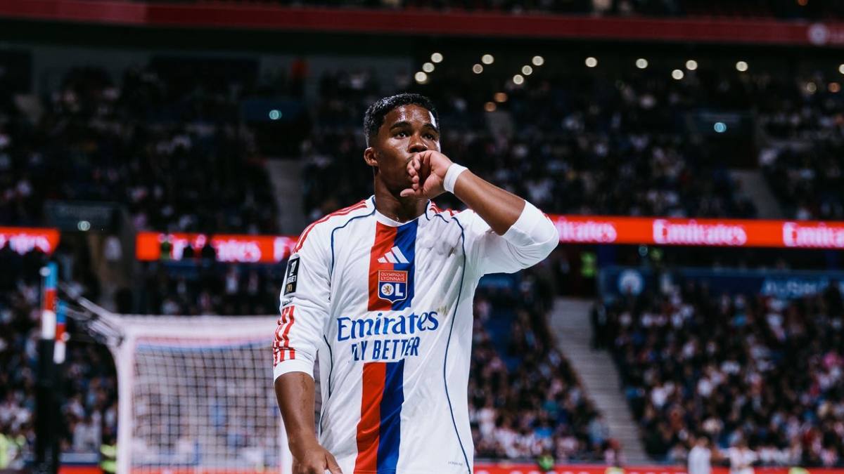 Endrick Lyon em partida no estádio, com uniforme do Olympique Lyon e o gesto de mão no rosto, em cena de torcedores ao fundo.