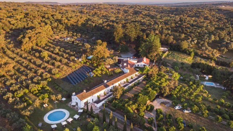 Aerial da Imani Country House no Alentejo, perto de Évora, cercada por campos e oliveiras, com casa de campo e piscina ao pôr do sol