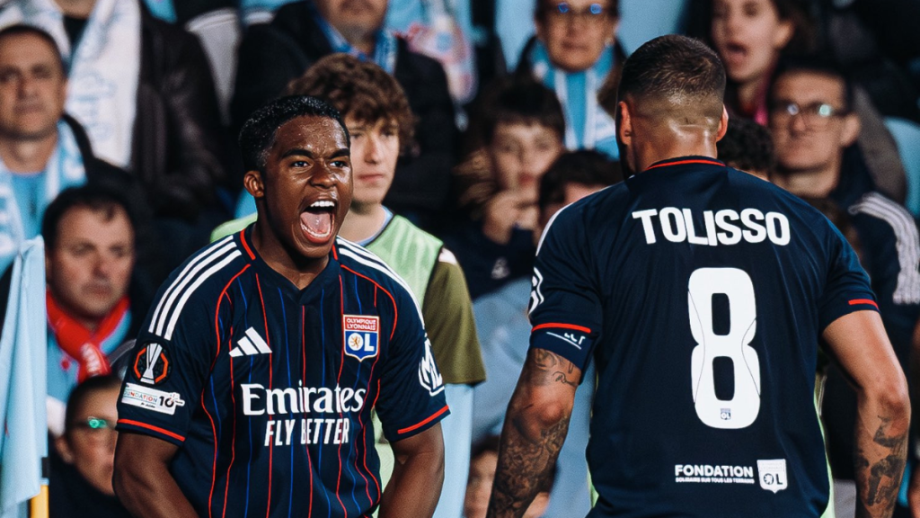 Endrick celebra gol pelo Lyon em partida com o uniforme do clube, comemorando ao lado de Tolisso, enquanto torcedores assistem ao fundo no estádio.