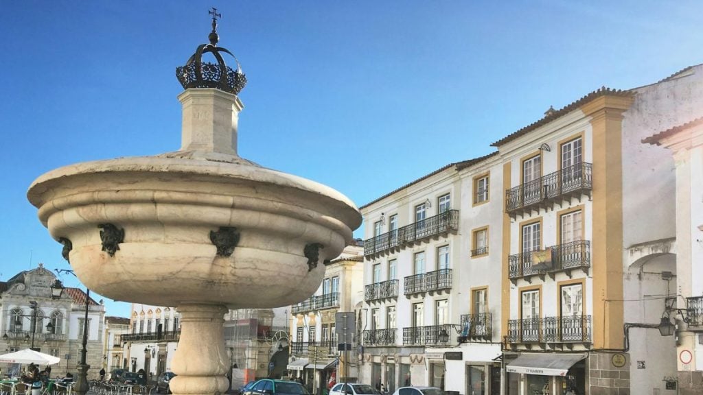 Imani Country House no Alentejo, perto de Évora: fachada histórica com fonte de pedra e edifícios amarelos e brancos com varandas ao ar livre sob céu azul.
