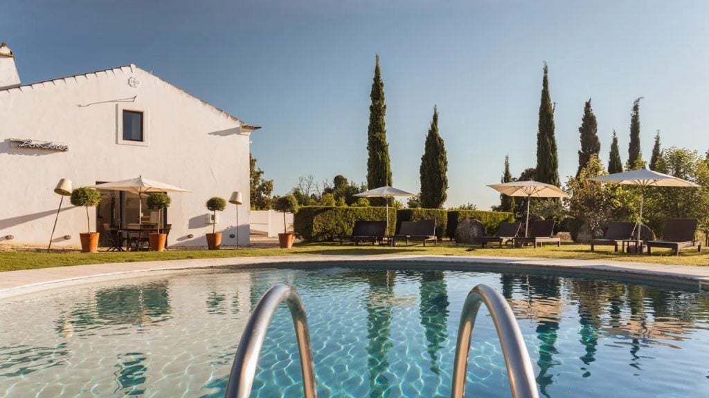 Imani Country House no Alentejo, perto de Évora: piscina ao ar livre com espreguiçadeiras e guarda-sóis ao lado de um edifício branco, jardim e céu azul.