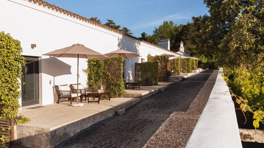Imani Country House no Alentejo, perto de Évora: pátio externo com cadeiras e mesas sob guarda-sóis, fachada branca e caminho de pedra entre jardins.