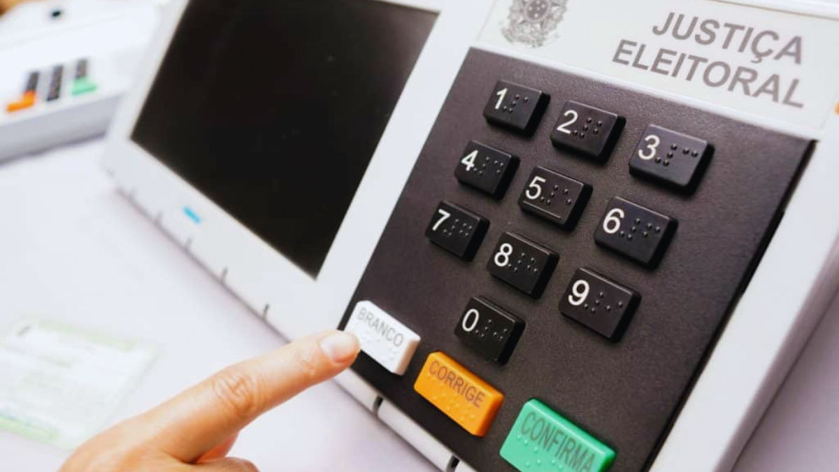 Eleições 2026 com urna eletrônica e identificação de Justiça Eleitoral, mostrando teclado numérico e botão de confirmação com a mão do eleitor prestes a votar