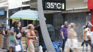 Mudanças bruscas de temperatura: pessoas circulam em rua comercial com painel digital marcando 35°C e variação térmica ao fundo, sugerindo “gangorra” climática em São Paulo