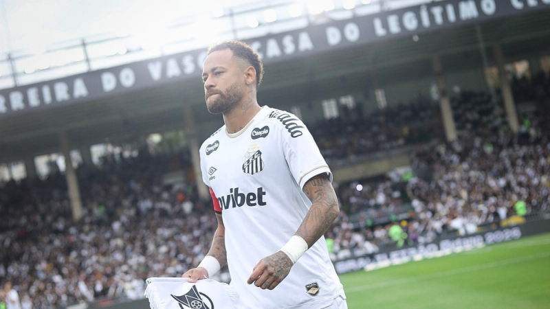 Neymar em estádio usando camisa do Santos, com expressão pensativa antes de partida; tema da imagem indica Neymar fora do clássico contra o Palmeiras e contra gramado sintético.