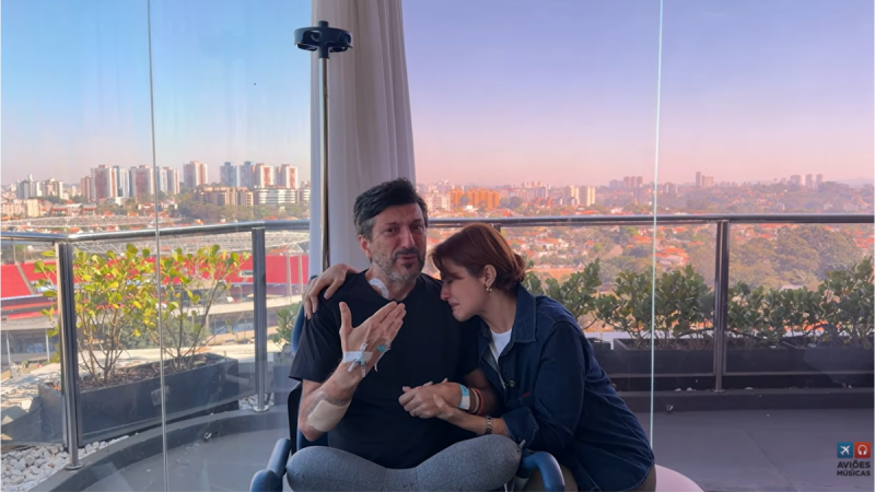 Casal sentado em uma área com grandes janelas, conversando e demonstrando carinho, enquanto ao fundo aparece uma vista panorâmica da cidade ao entardecer.