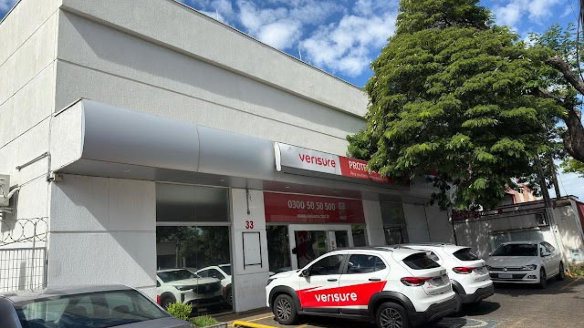Fachada de um imóvel comercial com cobertura, pátio de estacionamento e veículos estacionados, com identificação da empresa Verisure na entrada.