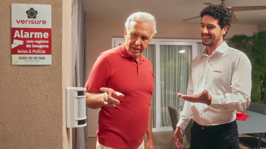 Instalação de sistema de alarme Verisure: técnico sorridente e cliente na entrada de uma residência observando o equipamento externo e o painel “Alarme com registro de imagens”.