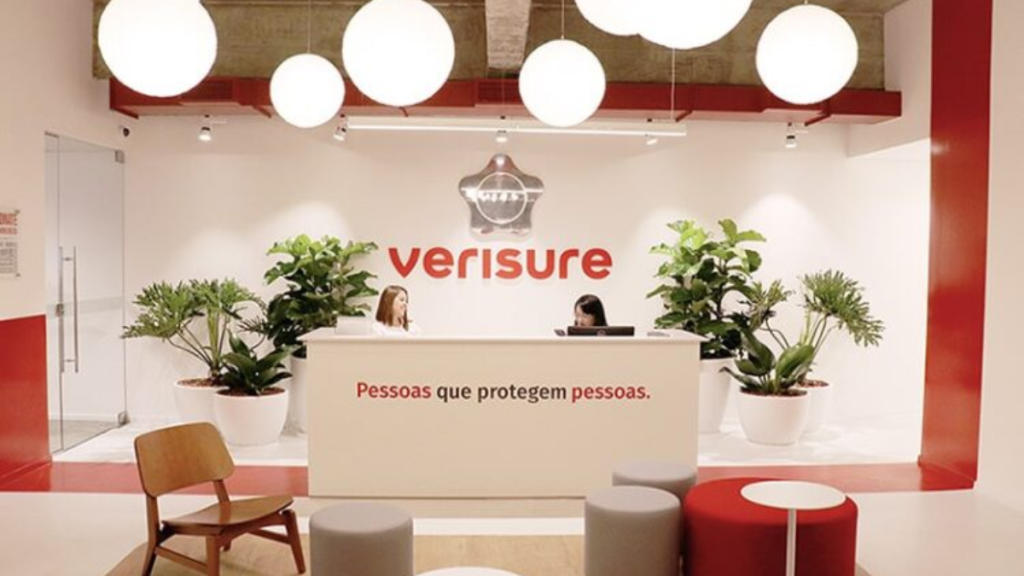Recepção corporativa da Verisure com balcão branco, frase “Pessoas que protegem pessoas.” e equipe ao fundo, além de plantas ornamentais e decoração moderna com iluminação pendente.