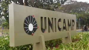 Unicamp 2027: placa com o logo e a sigla “Unicamp” em ambiente externo, com área verde ao fundo, relacionada ao vestibular que acontece em outubro.
