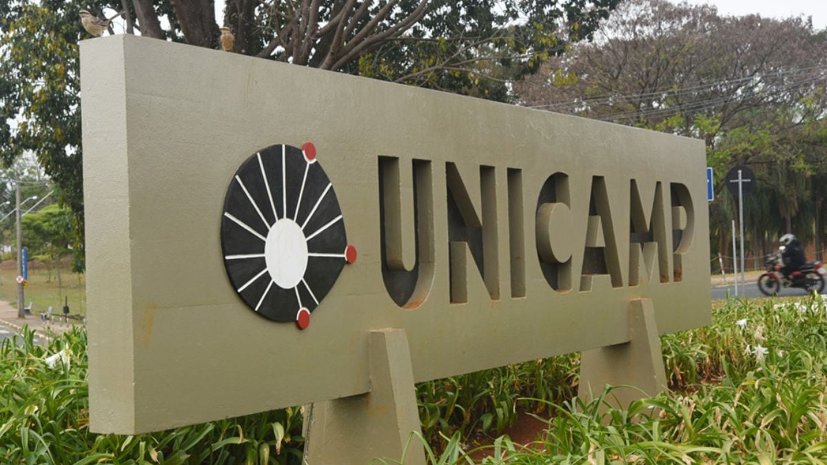 Unicamp 2027: placa com o logo e a sigla “Unicamp” em ambiente externo, com área verde ao fundo, relacionada ao vestibular que acontece em outubro.