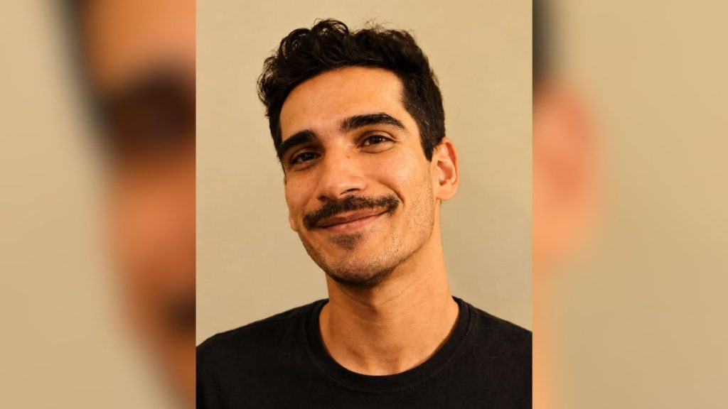 Vinícius é um jovem de pele clara e cabelos escuros, ligeiramente ondulados e curtos nas laterais. Ele possui sobrancelhas grossas, olhos escuros, um bigode aparado e uma leve barba rala no queixo. Veste uma camiseta preta de gola careca, e o fundo é liso, em tom bege neutro, com as bordas laterais levemente desfocadas. Ele sorri suavemente para a câmera. O jovem está desaparecido há 21 dias.