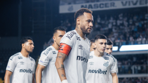 Neymar em campo durante partida do Santos, com uniforme branco e faixa vermelha no braço, em um estádio com torcedores ao fundo.