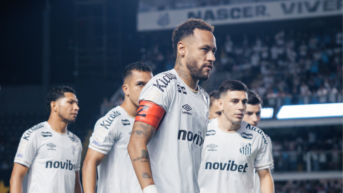 Neymar em campo durante partida do Santos, com uniforme branco e faixa vermelha no braço, em um estádio com torcedores ao fundo.