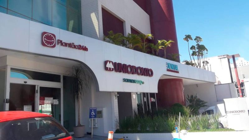 Fachada da clínica UniOdonto Campinas, com fachada branca, placa com o nome da unidade e entrada de atendimento, em ambiente urbano externo.